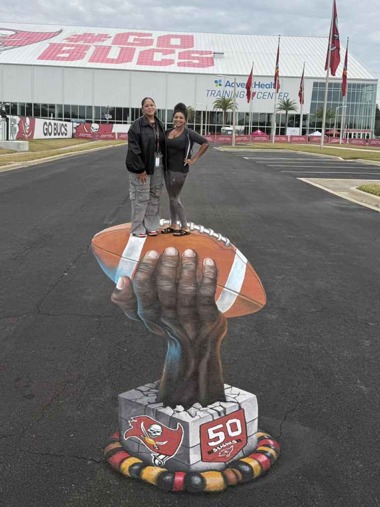 A wide shot at the Tampa Bay Buccaneers training facility. In the foreground, two people pose on a 3D street art painting that creates the illusion of a giant hand lifting a football out of the pavement. The base of the mural features the Buccaneers logo and the number 50. In the background, a large white building features "GO BUCS" in red lettering and several team flags.