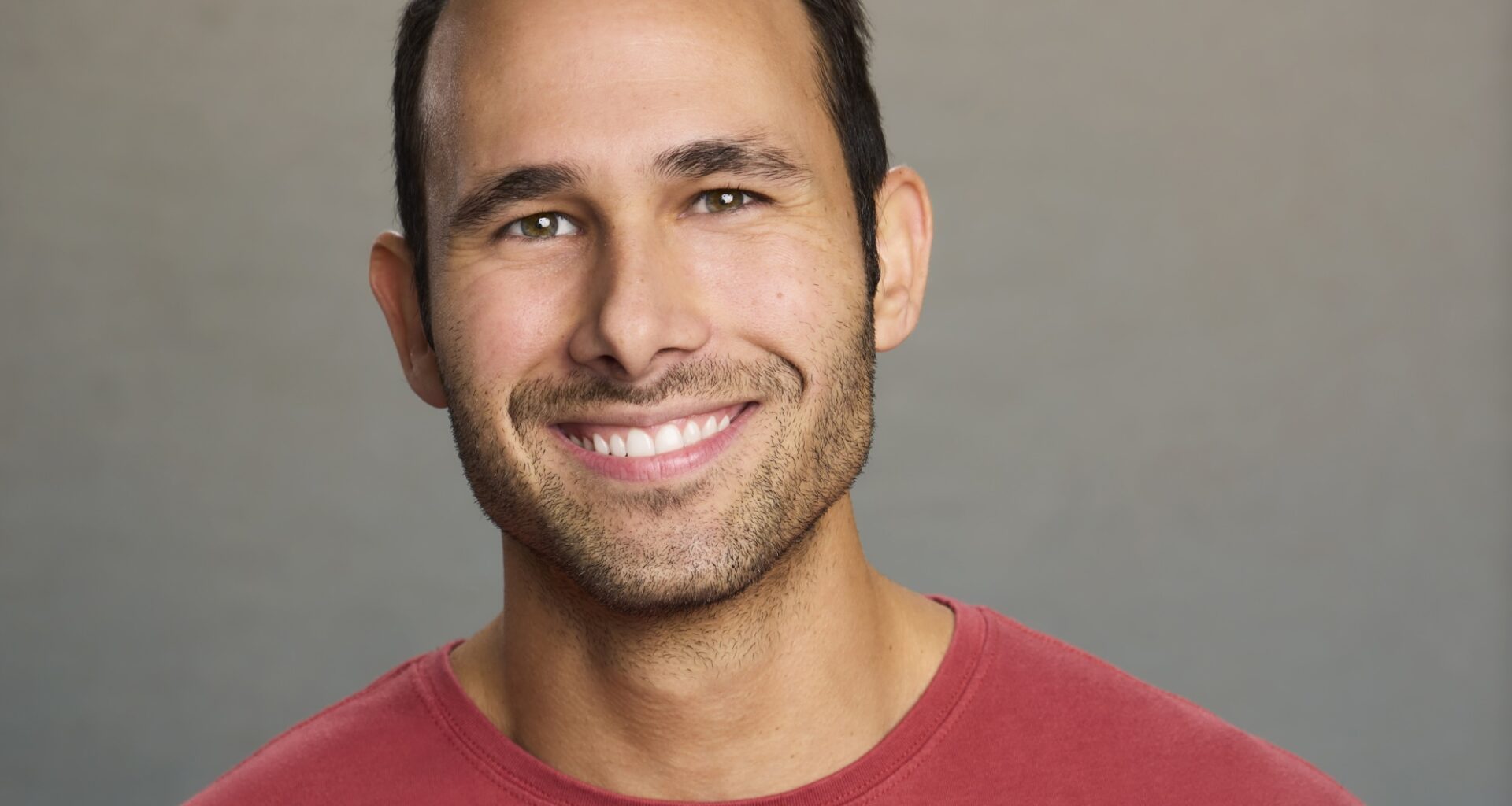 headshot of a smiling man wearing a red shirt in front of a gray background