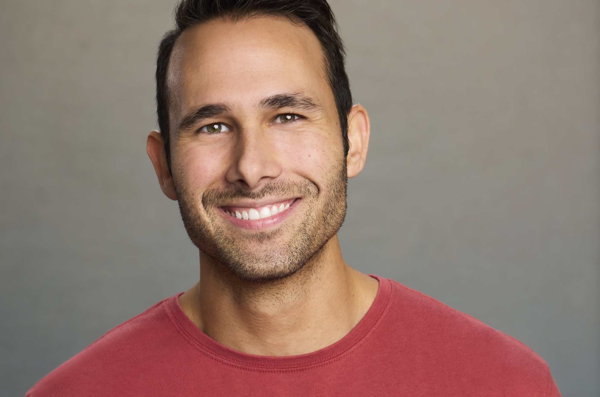 headshot of a smiling man wearing a red shirt in front of a gray background