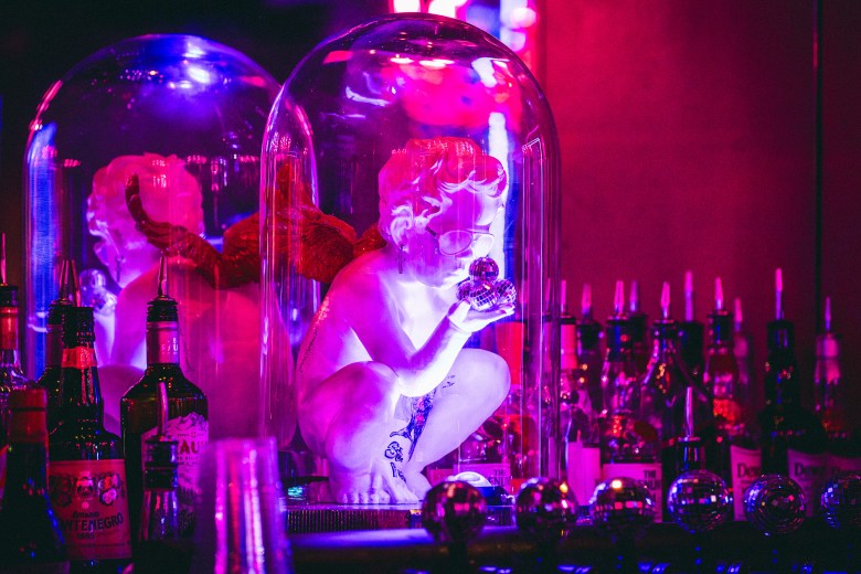 A white cherub figurine wearing sunglasses encased in a glass dome on a shelf behind a bar, illuminated by pink and purple lighting.