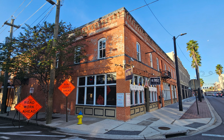 A wide-angle street view of a two-story historic red brick building on a corner, identified by a black sign as "THE BRICKS." The sidewalk in front features a yellow fire hydrant and two bright orange diamond-shaped construction signs that read "ROAD WORK AHEAD" and "ROAD CLOSED AHEAD." The scene is captured on a clear day with a blue sky and tall palm trees visible further down the street.