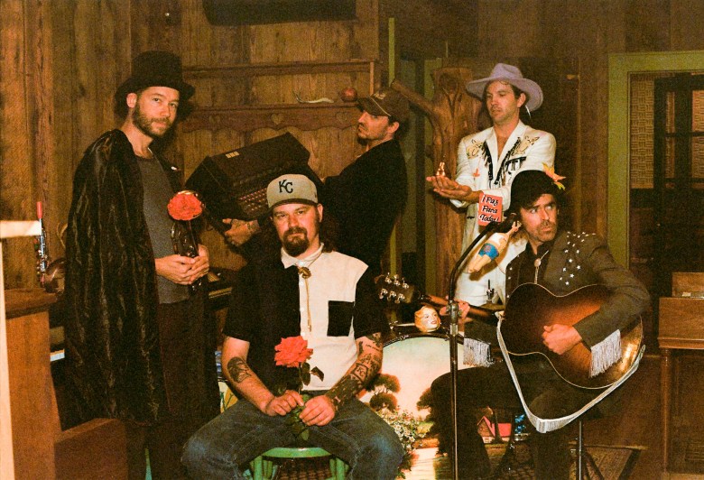 The Deslondes band members pose for a portrait by Selwyn Figg in a rustic, wood-paneled room. The five musicians are dressed in vintage-style Americana clothing, standing and sitting together in an informal arrangement that reflects their country-soul aesthetic.