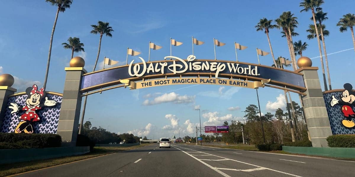 Entrance to Walt Disney World featuring a large archway with "The Most Magical Place on Earth" written below. Mickey and Minnie Mouse are displayed on pillars. Palm trees line the road under a blue sky.