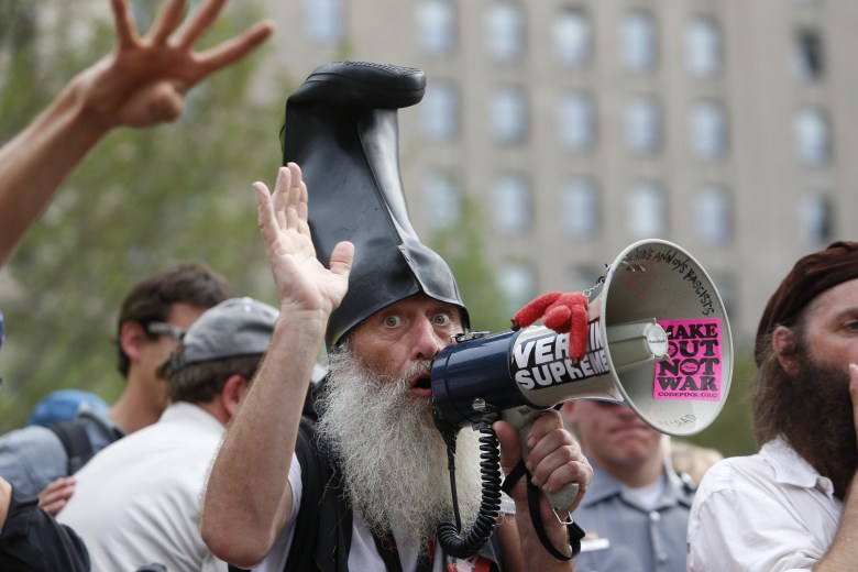 A person with a long, bushy white beard and wide eyes speaks into a megaphone. They are wearing a large black rubber boot on their head like a tall hat. The megaphone has a sticker that reads "VERMIN SUPREME" and another pink sticker that says "MAKE OUT NOT WAR." The individual has one hand raised, and the background shows a crowd of people in front of a tall, multi-story building.