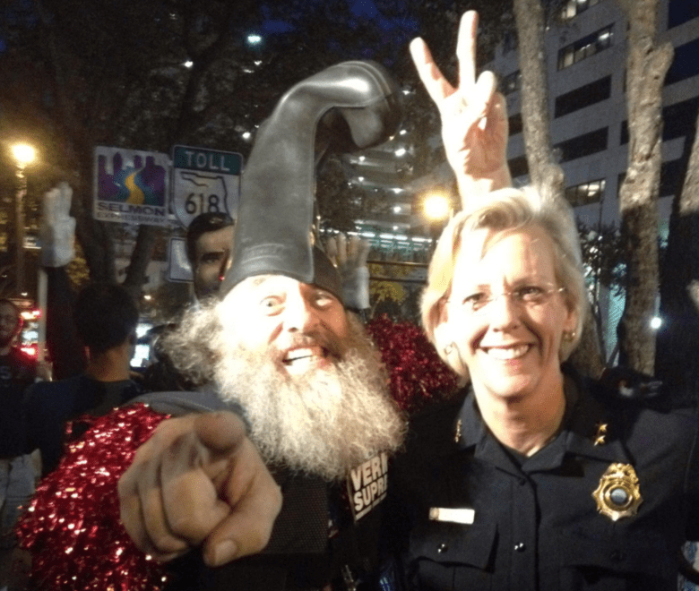 A night-time candid photo of two people smiling. On the left, a person with a long grey beard wears a large black rubber boot as a hat and a red sequined garment, pointing directly at the camera. On the right, a person in a dark blue police uniform with a gold badge smiles while holding up a peace sign with their hand. The background shows street signs for the "Selmon Expressway" and "TOLL 618" among city trees and buildings.