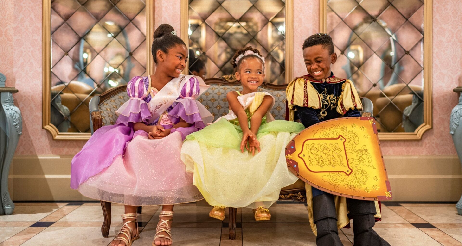 Three smiling kids in princess and knight costumes sit on a bench, echoing the royal flair of a Disney Park&rsquo;s castle interior.