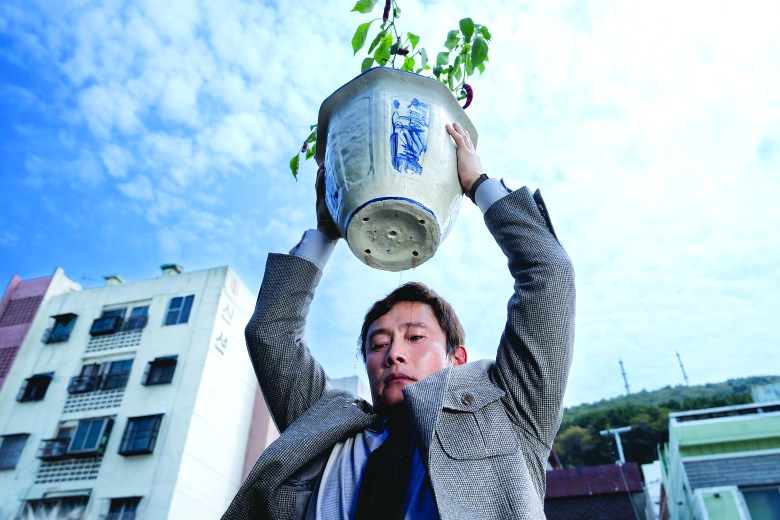 A low-angle shot of a man (actor Lee Byung-hun) wearing a grey suit jacket. He holds a large, white-and-blue ceramic pot containing a pepper plant directly above his head. Water drips from the bottom of the pot. In the background, a white apartment building with Korean text is visible against a cloudy blue sky