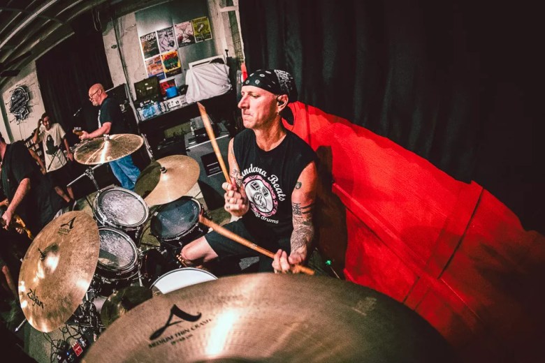 rummer Shawn Watkins of the band Big Sad performing live; a wide-angle shot showing him behind a drum kit with a black bandana, mid-swing with drumsticks in a rehearsal or venue space.
