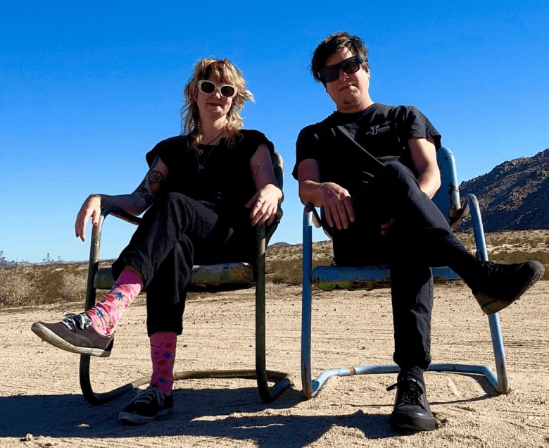 Two people sit side-by-side in weathered metal chairs in a vast, sandy desert landscape under a clear, bright blue sky. Both are wearing black t-shirts and black pants. The individual on the left has blonde hair, white-framed sunglasses, and bright pink patterned socks. The individual on the right is wearing dark sunglasses and has their legs crossed. A rocky hill is visible in the distant background.