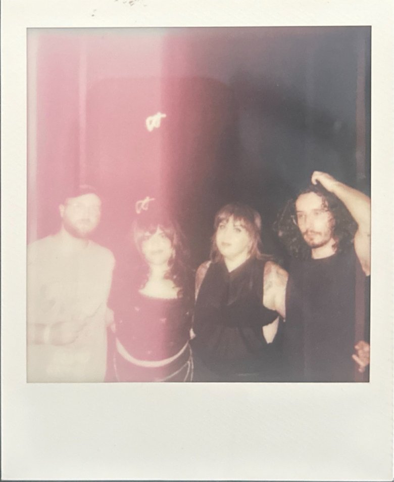 slightly overexposed, Polaroid-style photograph of four people standing together in a dimly lit indoor space. From left to right: a person in a baseball cap and light-colored t-shirt, a person with long dark hair wearing a dark patterned top, a person with shoulder-length brown hair and visible arm tattoos in a black sleeveless top, and a person with long curly dark hair wearing a black t-shirt with one hand resting on their head. The image has a soft, nostalgic aesthetic with a prominent hazy red light flare on the left side.