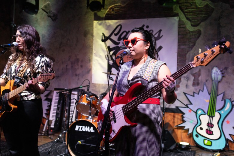 Two women perform on a stage with a brick wall background. On the left, a guitarist in a shiny gold animal-print top plays an electric guitar. On the right, a bassist wears red heart-shaped sunglasses and a checkered jumpsuit while playing a red bass guitar. Behind them is a drum kit and a banner with the text 'Ladies Rock.'