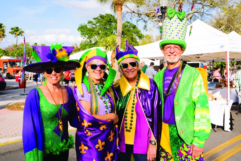 A cheerful group of four older adults dressed in colorful purple, green, and gold Mardi Gras costumes at an outdoor festival.