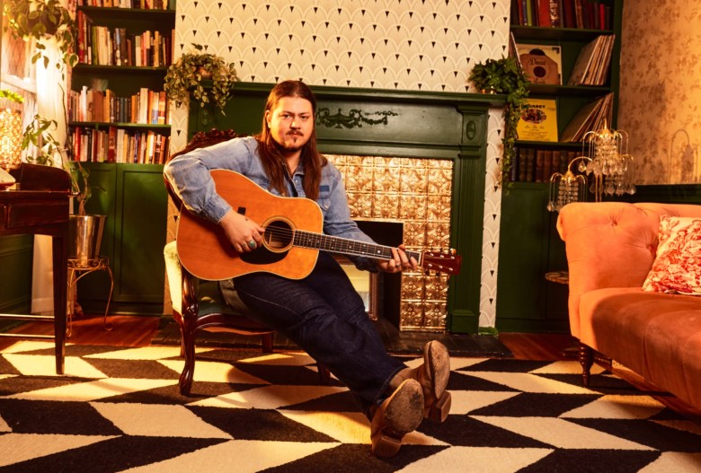 Vincent Neil Emerson, wearing a denim shirt and jeans, sits on a wooden chair and plays an acoustic guitar. He is in a cozy room with dark green bookshelves flanking a fireplace, a patterned black and white wall, and a large black and white checkered rug. A pinkish-orange velvet couch is visible on the right.