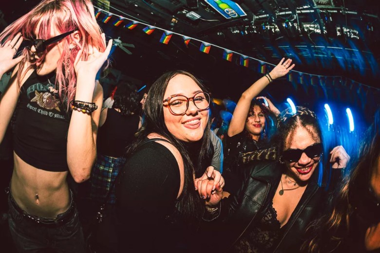 A woman wearing glasses smiles while holding hands and dancing with another woman, other woman can be seen dancing in the background in front of a stage