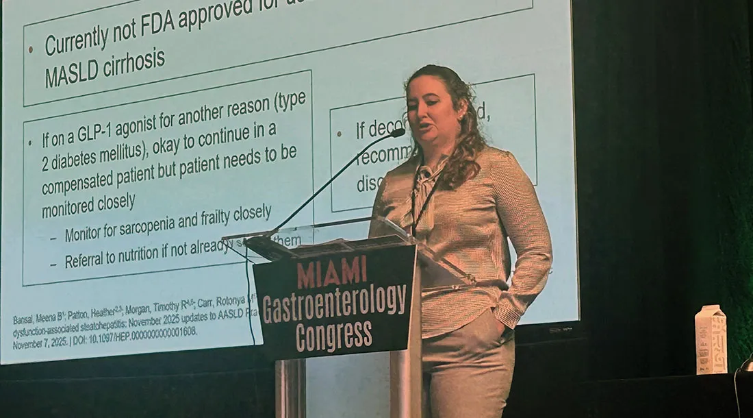 Dr. Lauren Gilbert stands at a podium labeled “Miami Gastroenterology Congress,” delivering a talk in front of a large projected slide with clinical bullet points about MASLD cirrhosis and GLP‑1 agonist use, in a darkened conference setting.