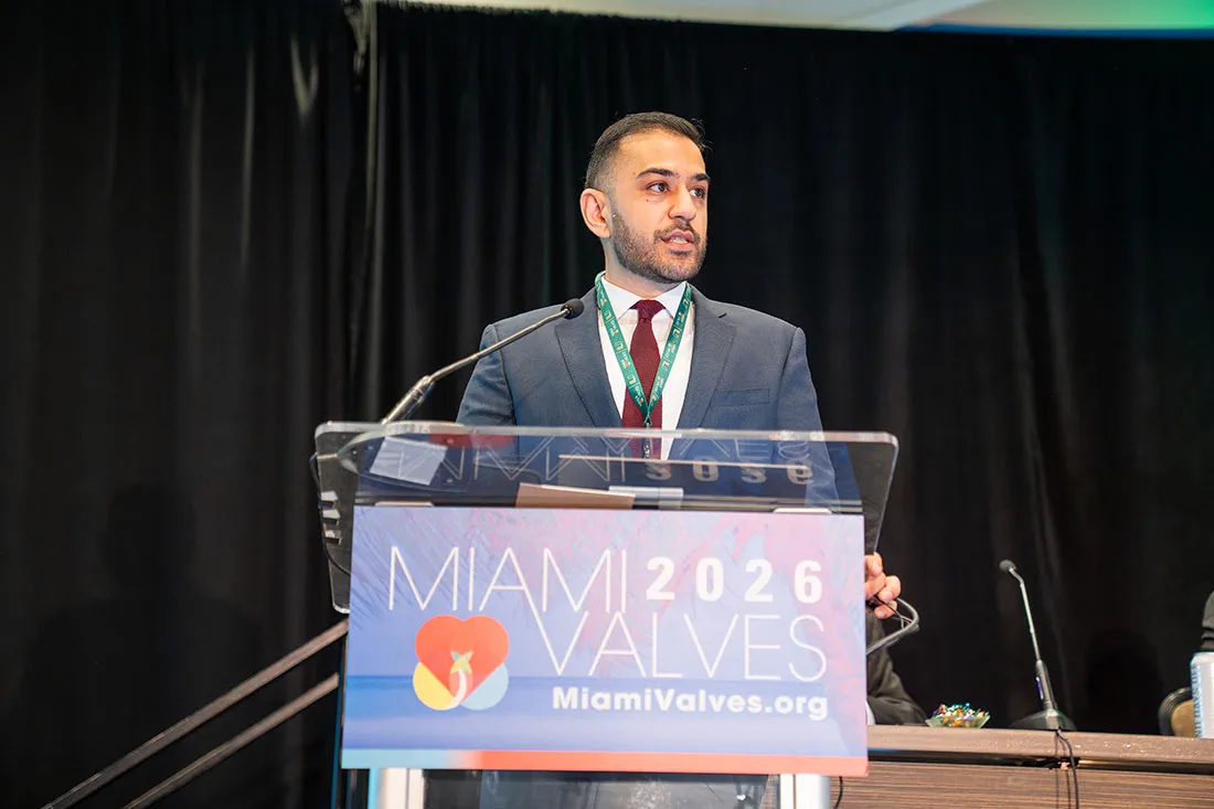 Dr. Carlos Collet in a dark suit stands at a clear podium on a stage, addressing an audience at the Miami Valves 2026 conference. The podium displays the event’s colorful logo and the website MiamiValves.org. Thick black curtains form the backdrop, and a microphone is positioned in front of the speaker.
