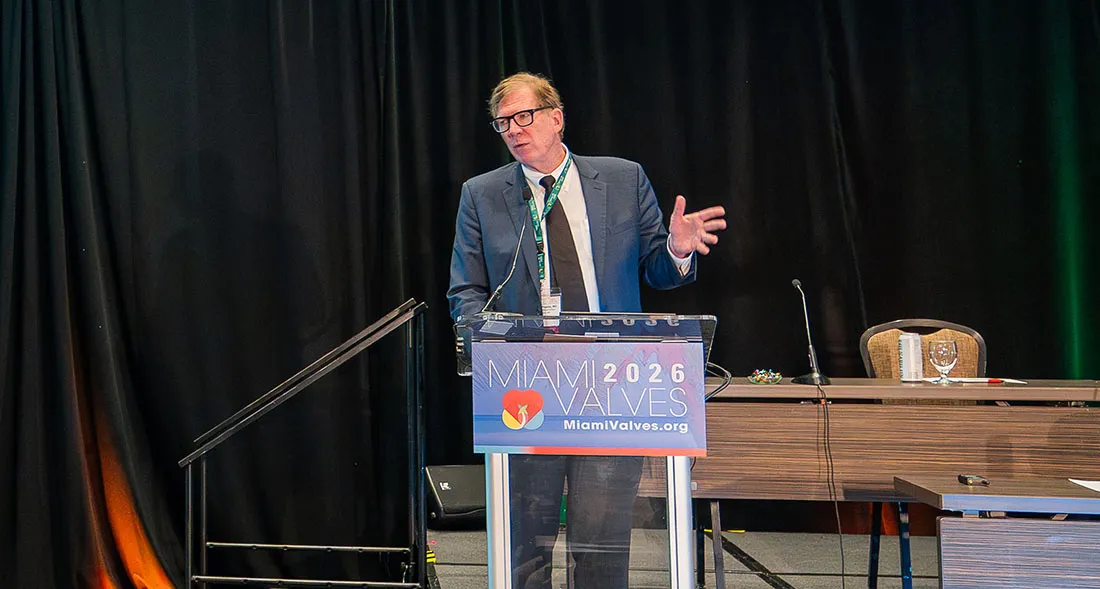 Dr. Jeffrey Popma in a suit stands at a clear podium on a conference stage, gesturing with one hand while addressing the audience. The podium displays signage for “Miami Valves 2026” with the event logo and website. A long table with microphones, a water glass, and chairs sits behind the podium against a dark curtain backdrop.