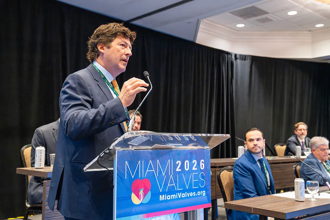 Dr. Juan Pablo Umaña stands at a clear podium branded with the “Miami Valves 2026” logo, addressing an audience in a conference room. Several seated panelists in suits are visible behind the podium. The backdrop features black curtains and overhead lighting typical of a professional medical meeting.
