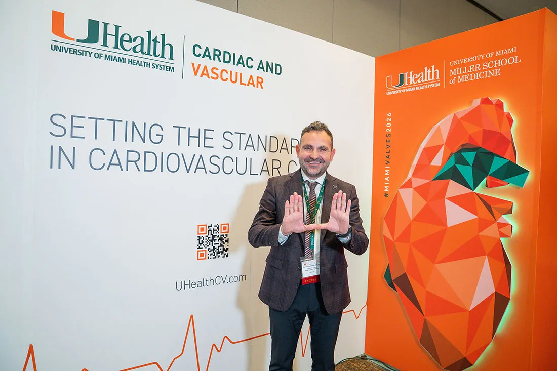 Dr. Yiannis Chatzizisis stands in front of a UHealth Cardiac and Vascular backdrop, holding up the University of Miami “U” hand gesture. The display includes the UHealth logo, a large geometric heart graphic, and the phrase “Setting the Standard in Cardiovascular Care.”