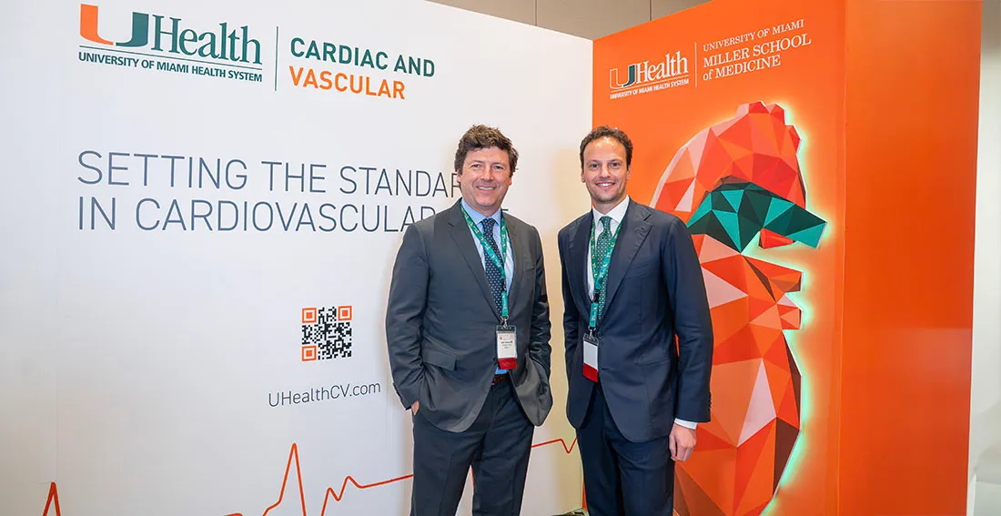 Dr. Juan Pablo Umaña and a colleague stand in front of a University of Miami Health System display at a cardiovascular event. Behind them, the backdrop features the UHealth logo, the words “Cardiac and Vascular,” and the slogan “Setting the Standard in Cardiovascular Care."
