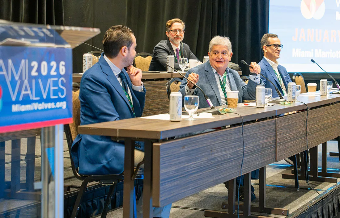 Dr. De Marchena and a panel of speakers in suits sits at a long conference table discussing a topic at the Miami Valves 2026 event. Microphones, water glasses, and event signage are visible, including a podium showing the Miami Valves 2026 logo.
