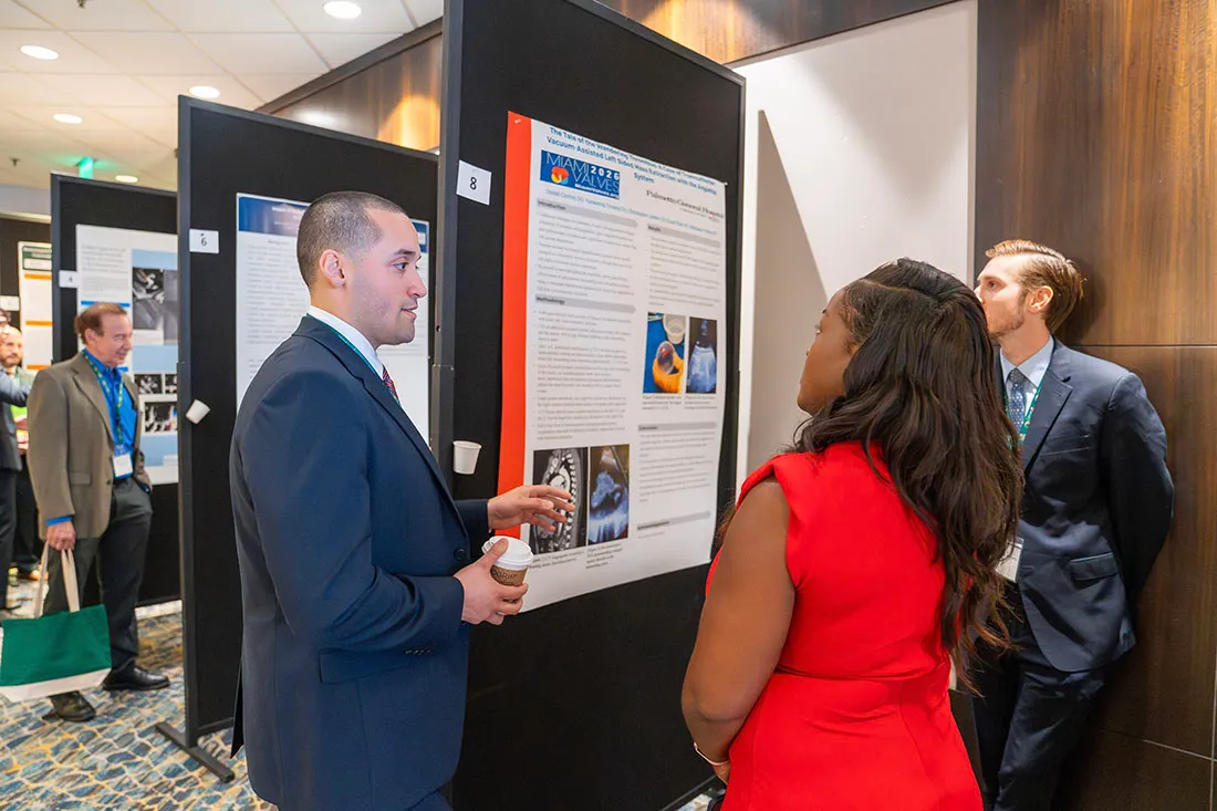 A small group of professionally dressed Miami Valves attendees stand in front of a large scientific poster at a conference. One person gestures toward the poster while holding a cup, as others listen and read the displayed research. Additional posters and attendees are visible in the background along a carpeted hallway.