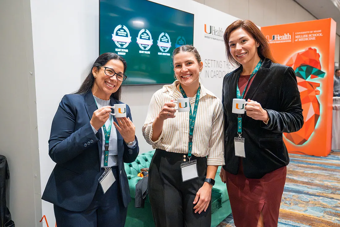 Three individuals in conference attire stand together holding white mugs with the University of Miami “U” logo. A UHealth Cardiac and Vascular display is visible behind them, along with a monitor showing award badges.