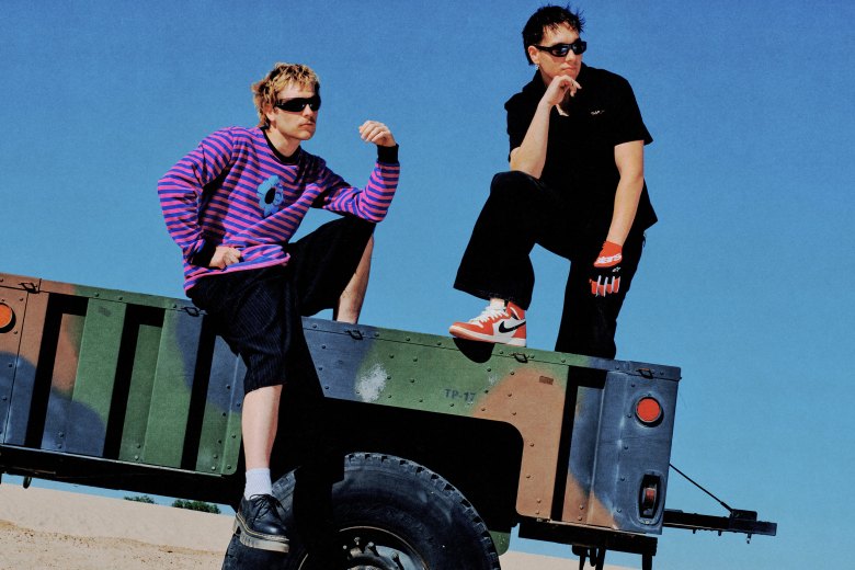 Joey Valence and Brae posing on a camouflaged military trailer in a desert setting. Joey Valence (right) stands in an all-black outfit with orange Nike Dunk high-tops and a red Alpinestars glove, while Brae (left) sits wearing a pink and blue striped "Golf Wang" style flower shirt and black shorts. Both artists are sporting their signature 90s-inspired streetwear and wraparound sunglasses against a clear blue sky.