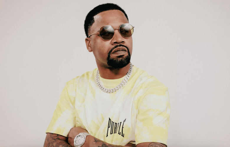 New Orleans rapper Juvenile posing in a yellow and white tie-dye t-shirt, wearing round gold-rimmed sunglasses, a thick silver Cuban link chain, and a luxury watch with his arms crossed against a plain white background.