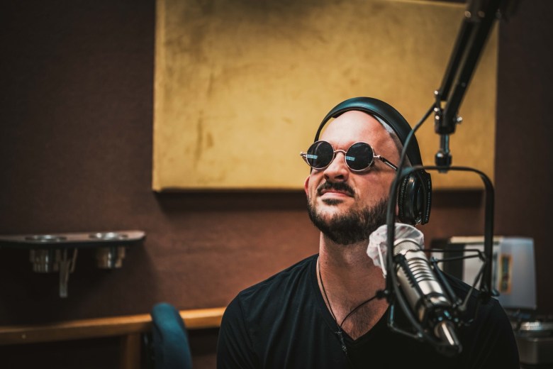 Tampa Bay musician Ken Apperson in a radio studio, wearing small round sunglasses and headphones while speaking or singing into a professional broadcast microphone. The warm-toned studio background features acoustic foam and a gold-colored sound panel.