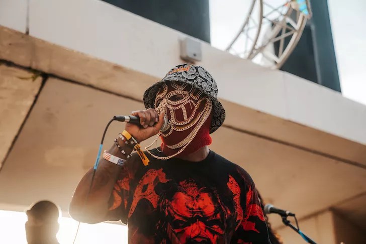 ampa hip-hop artist Pusha Preme performing on stage wearing a signature gold chain face mask, a patterned bucket hat, and a red and black graphic shirt.