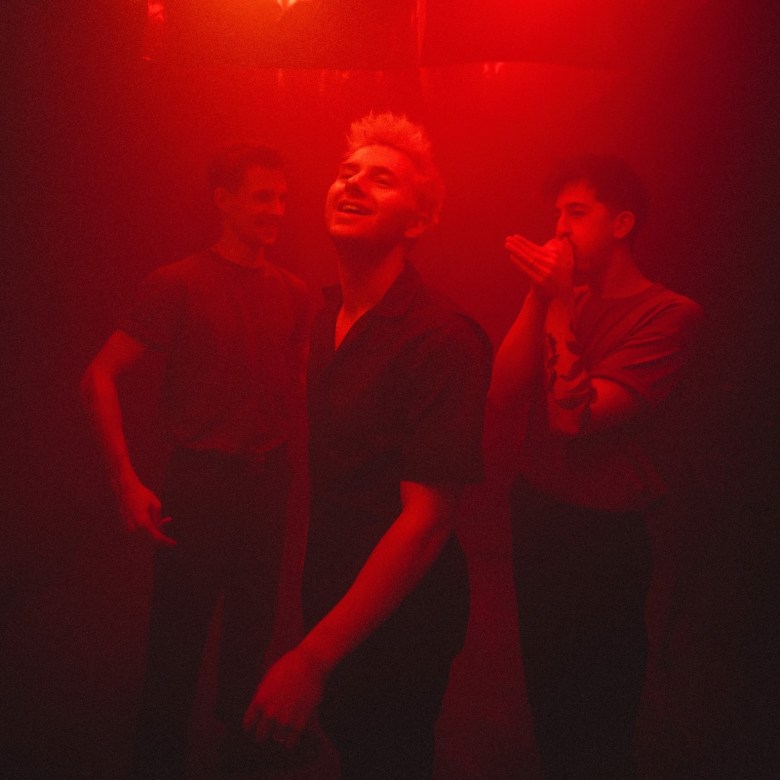Three people are captured in a candid moment under heavy, atmospheric red lighting and a slight haze. In the center foreground, a person with short, light-colored hair looks upward with a joyous expression, wearing a dark short-sleeved button-down shirt. Behind them to the left, another person smiles while looking toward the center, and to the right, a third person with a visible arm tattoo holds their hands up near their face.