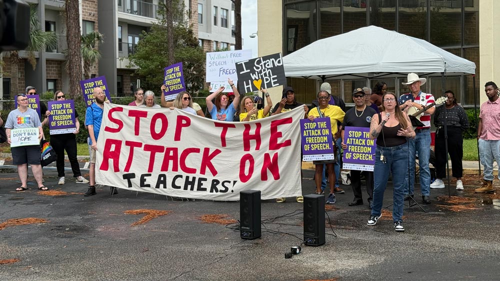 Protesters stand in front of DCPS headquarters holding a large banner that reads "STOP THE ATTACK ON TEACHERS" in bold, handwritten letters. The protest was in support of teachers' right to free speech.