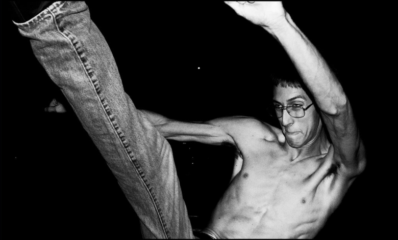 A dramatic black-and-white, high-contrast action shot of a shirtless person with short hair and glasses. They are captured mid-movement with one leg kicked high into the air toward the left of the frame and one arm raised, their muscles tensed and expression focused. The background is entirely black, making the subject appear spotlighted in the darkness.