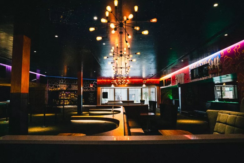Interior view of the dimly lit Sun-Ray Cinema lounge and bar area in Tampa. Industrial-style sputnik chandeliers with exposed amber bulbs hang from a dark ceiling over curved booths, with colorful neon accent lighting and mural art on the walls.