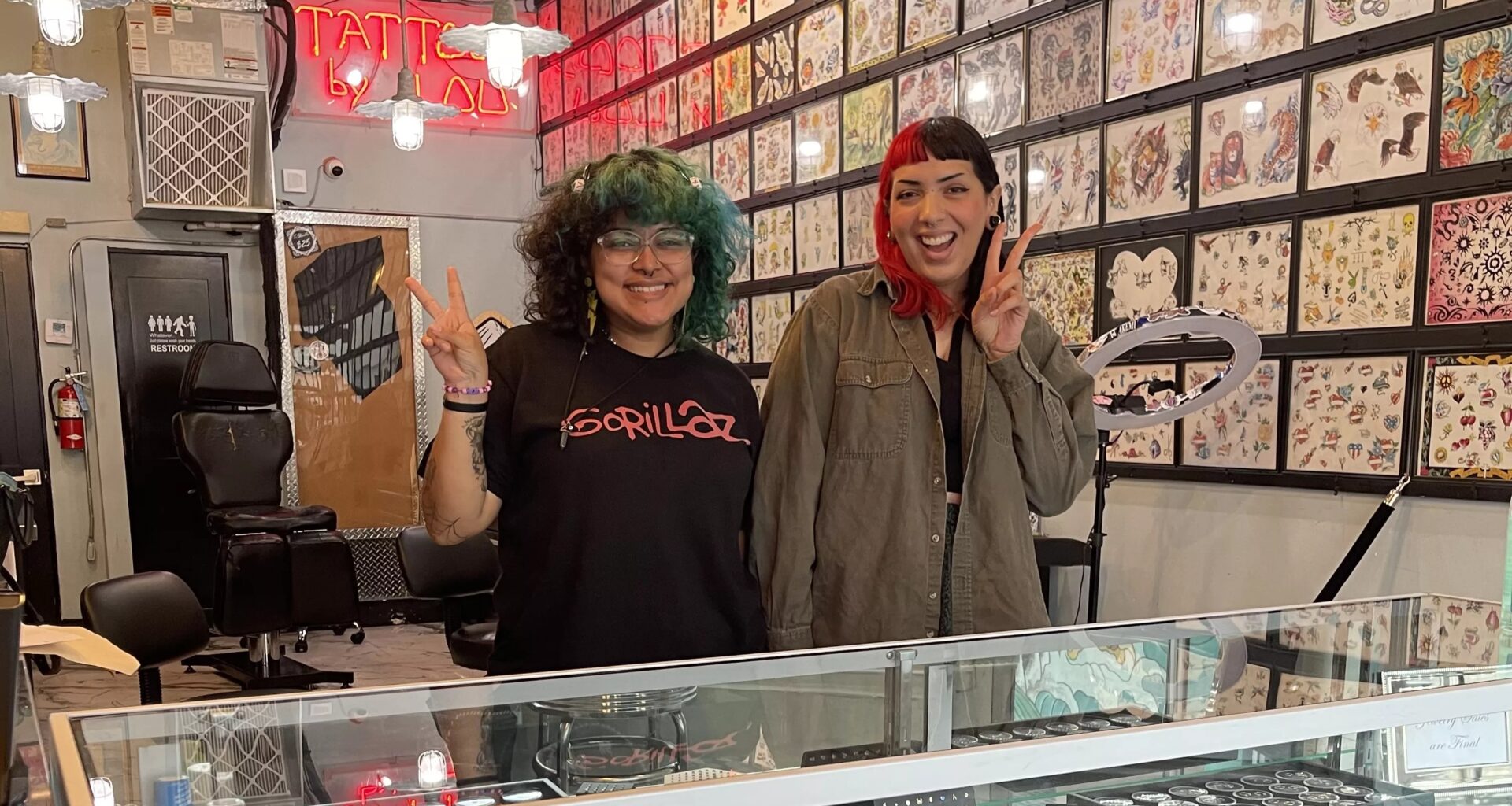 photo of two young women flashing the peace sign inside a tattoo studio with designs on the walls
