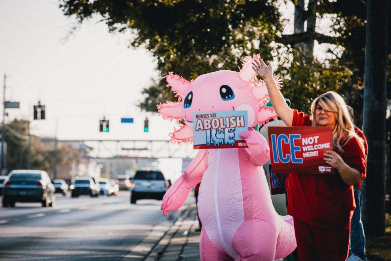 A person in an inflatable pink axolotl costume and a woman in red scrubs standing by a road holding signs that say "Nurses say abolish ICE" and "ICE not welcome here"