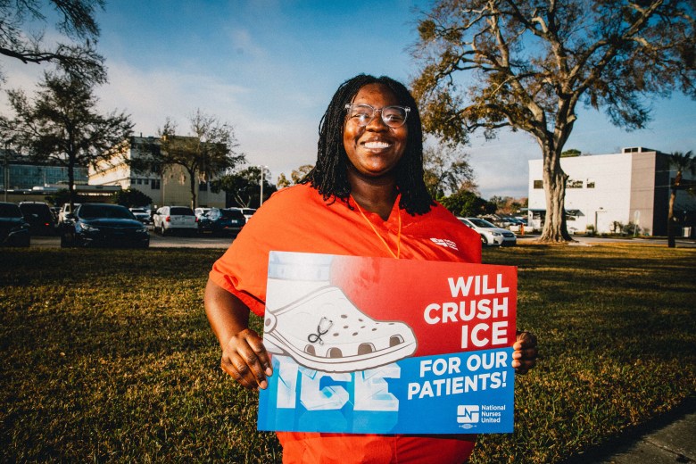 A smiling woman in orange scrubs holds a sign that reads “Will crush ICE for our patients in a grassy area outside a hospital.