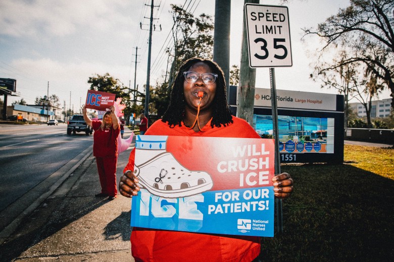 A smiling woman in orange scrubs with a whistle in her mouth holds a sign that reads “Will crush ICE for our patients in a grassy area outside a hospital.
