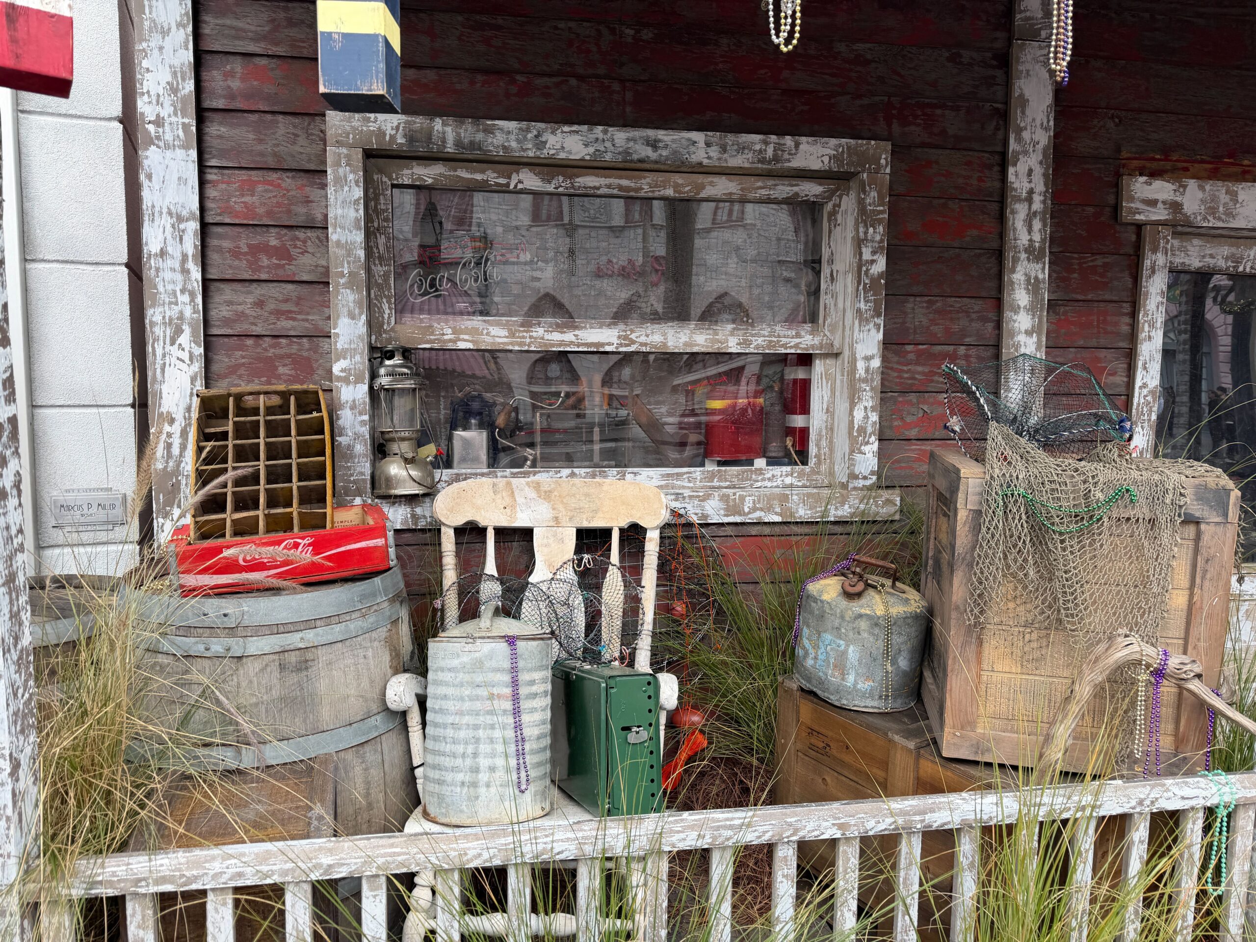 Crates, barrels, and a rocking chair sit on the porch. Decorations on the furniture include old Coca-Cola boxes, Mardi Gras beads, and fishing nets. More beads hang from the roof.