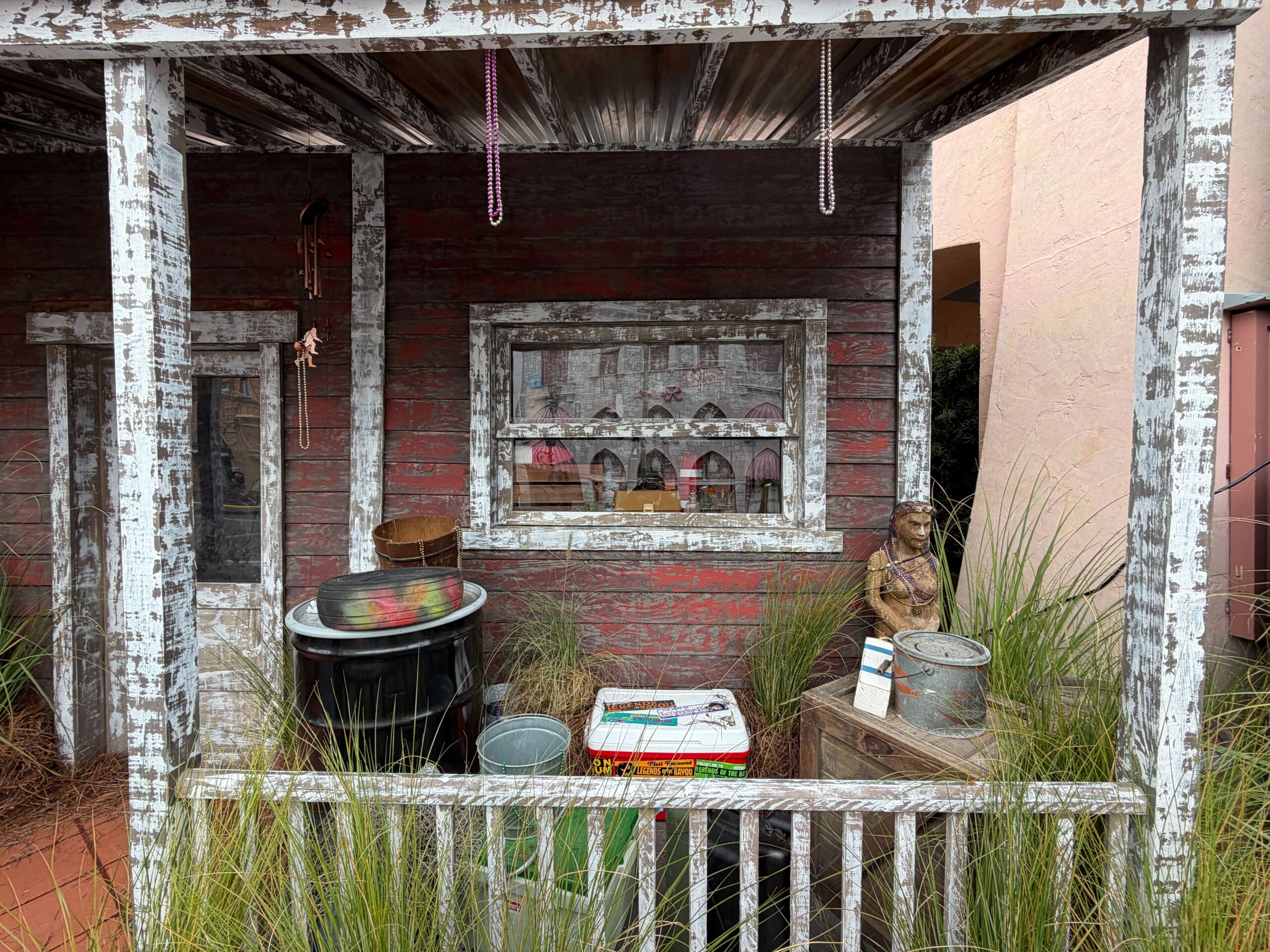 Crates, barrels, and a rocking chair sit on the porch. Decorations on the furniture include old Coca-Cola boxes, Mardi Gras beads, and fishing nets. More beads hang from the roof.