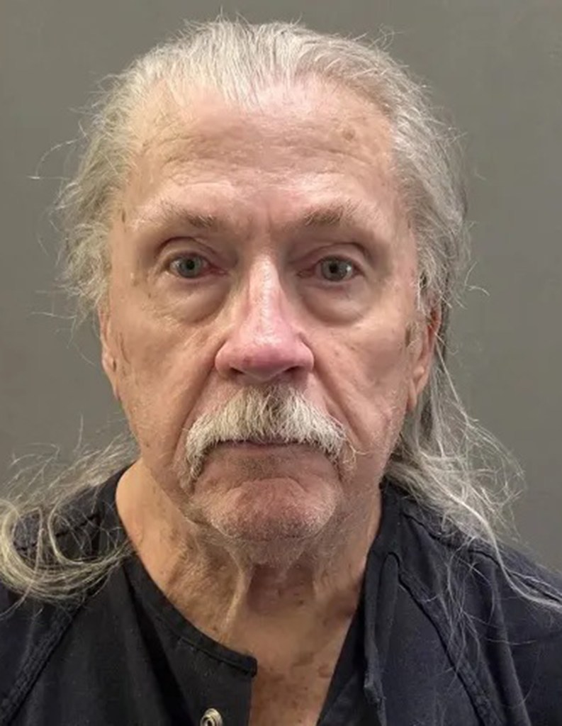 Mugshot of William Elwood Simmons, an 80-year-old white man with long gray hair and a mustache, wearing a black shirt.