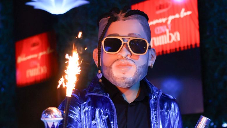 A person wearing an oversized caricature mask of the rapper Bad Bunny, complete with braided hair and sunglasses, wearing a shiny blue jacket and sunglasses holding a lit sparkler at a party.