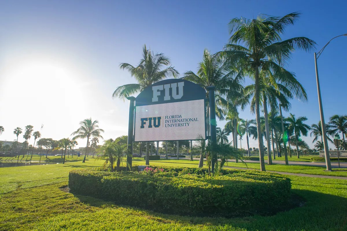 photo of a sign of Florida International University surrounded by lush grass, palm trees, and a clear blue sky