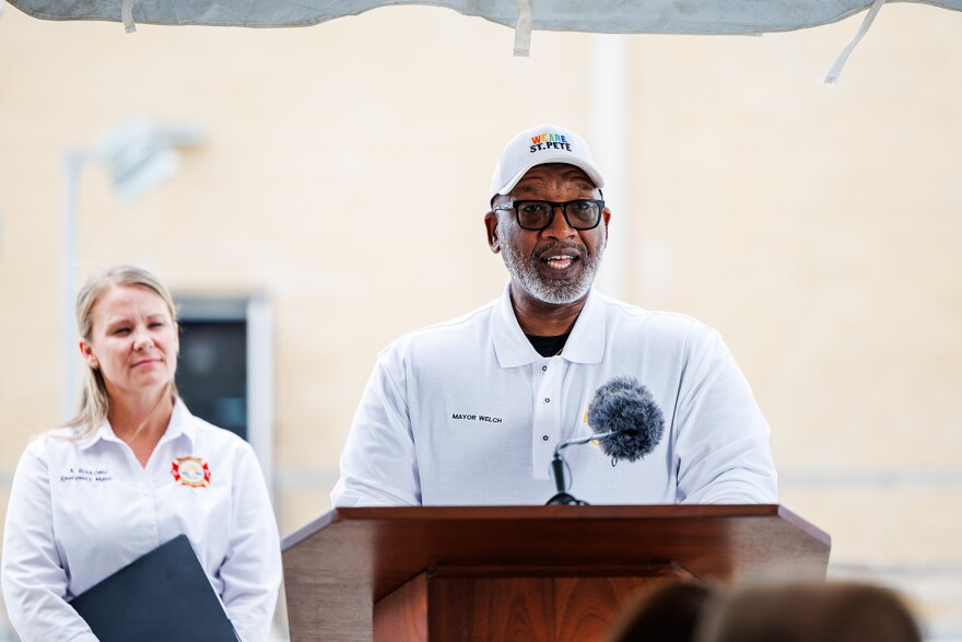 Man with a gray beard, white cap and polo shirt standing behind a podium with a blonde woman in a white button-down shirt to the left