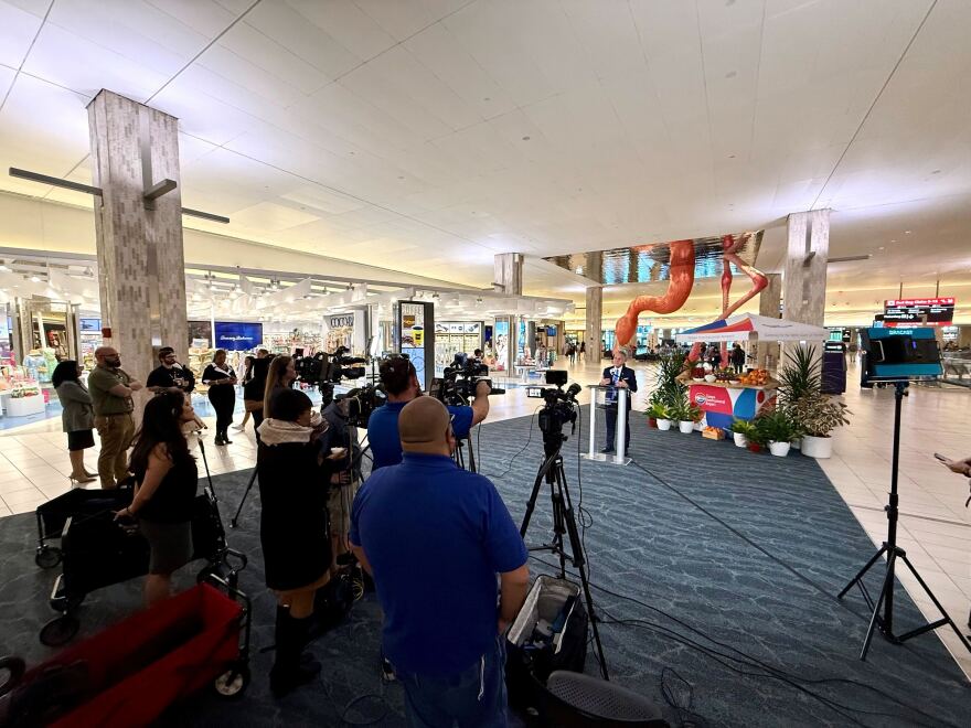 People with cameras record a man in a suit standing behind a podium in an airport