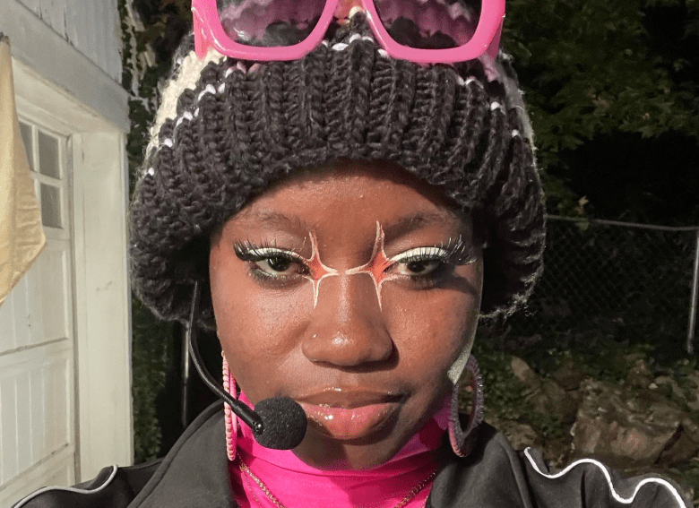 Musician Emmpowerr in a close-up portrait featuring artistic orange and white starburst eye makeup. The person wears a pink turtleneck under a black track jacket with white piping, standing outdoors at night.