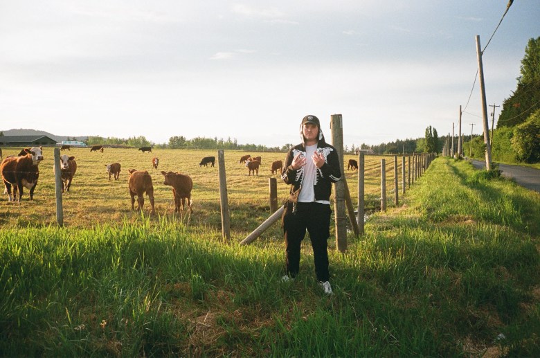 Musician Liam Hayden, known as Cult Member, standing in a grassy field at sunset. The person wears a black zip-up hoodie with a white skeleton print on the chest, black pants, and a black baseball cap. A long wooden fence and grazing cattle are visible in the background.