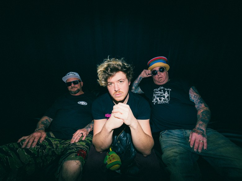 Members of the band Sublime, including Jakob Nowell, Eric Wilson, and Bud Gaugh, posing for a 2026 promotional photo. Jakob sits in the center with hands clasped, flanked by the original members against a dark, moody background.