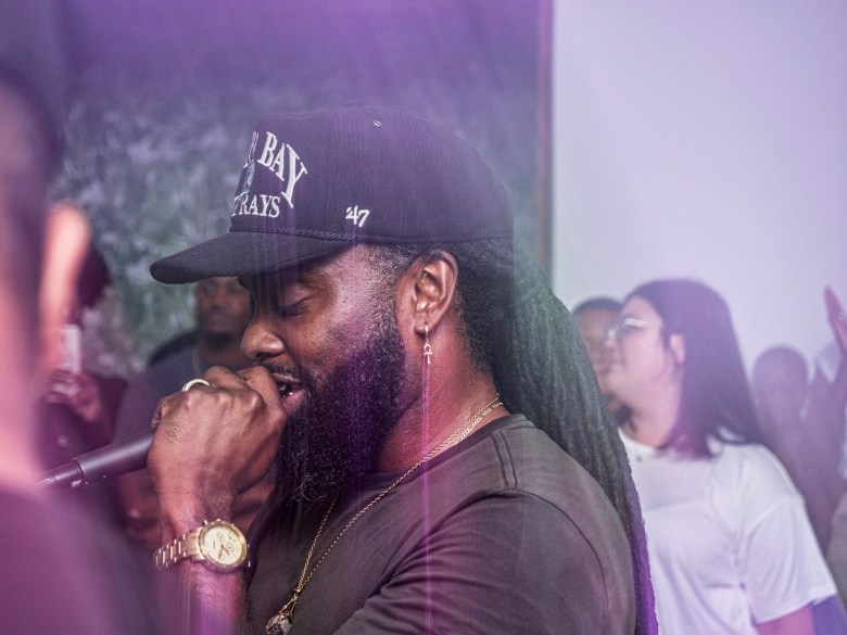 Musician J.IN.X performing live in a profile shot, wearing a black Tampa Bay Rays baseball cap and a gold watch while singing into a microphone. The person has a full beard and stands amidst stage smoke with a crowd visible in the blurred background.